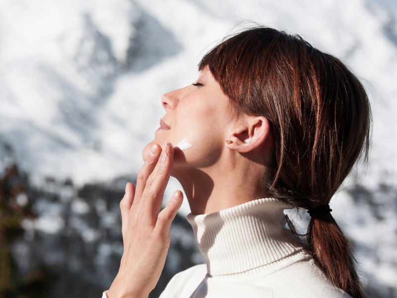 Woman with turtleneck sweater applies cream on her face and in the background there is snow on the mountains