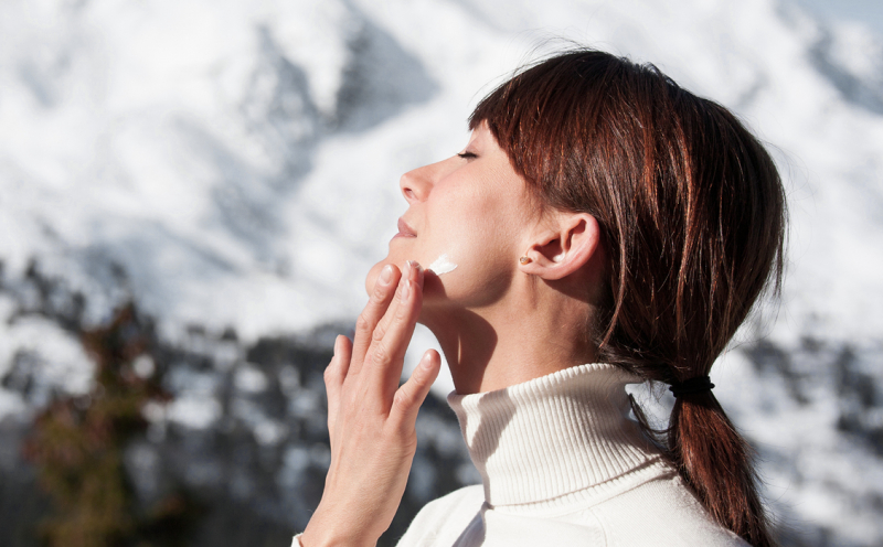 Woman with turtleneck sweater applies cream on her face and in the background there is snow on the mountains